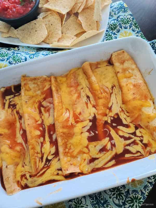 cheese enchiladas in a white baking dish next to a platter with chips and salsa