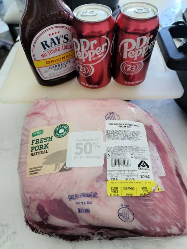 BBQ sauce, Dr Pepper cans, and packaged pork but on a white counter near a slow cooker