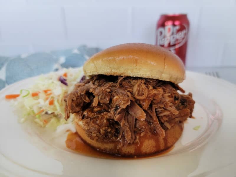 Pulled pork sandwich on a white plate with cole slaw near a can of Dr. Pepper 