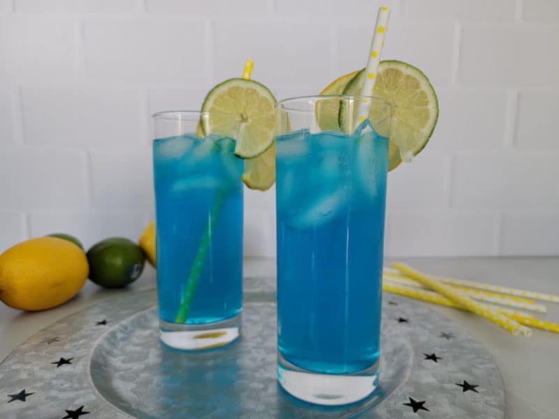 Two electric blue lemonade cocktails on a silver platter with yellow straws and lemon and limes