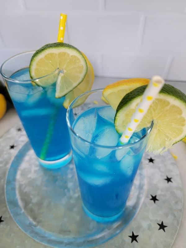 Two electric lemonade cocktails in tall glasses with paper straws and lemon and lime wheels for garnish