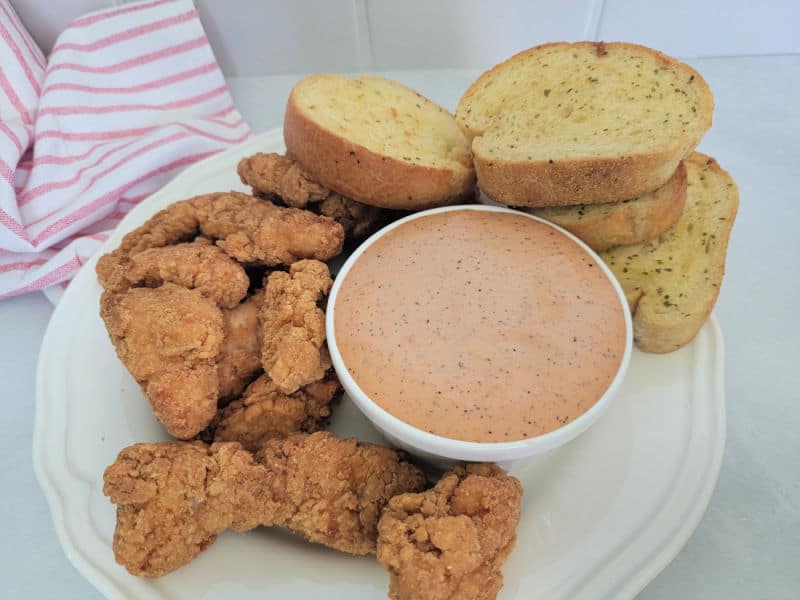 a white bowl filled with raising canes sauce on a white plate with garlic bread, and chicken pieces 