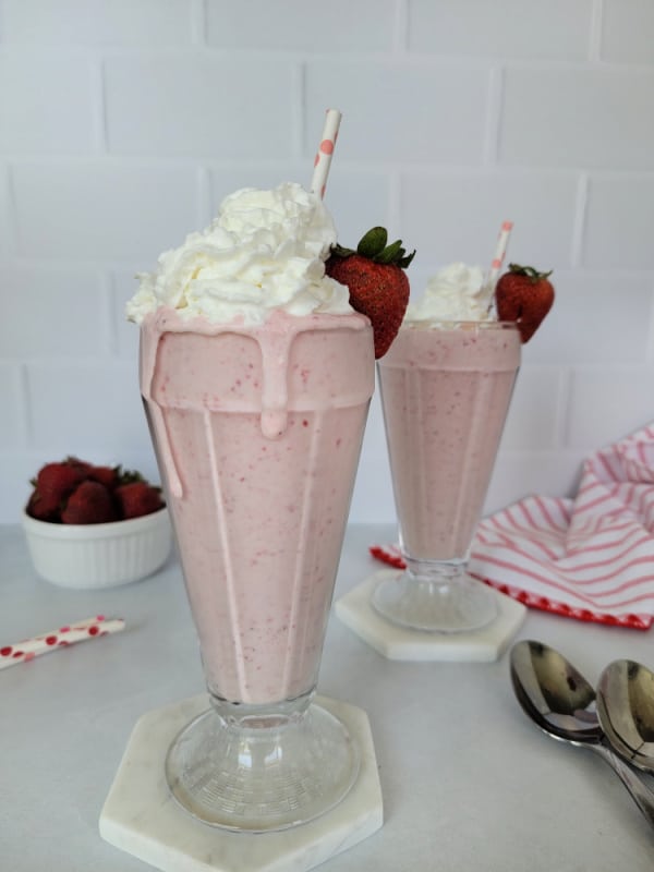 Two strawberry milkshakes in glasses with a bowl of strawberries, and a red striped cloth napkin