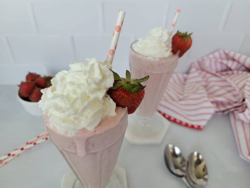 2 strawberry milkshakes with whipped cream garnish in glasses, bowl of strawberries, spoons, and a red striped cloth napkin