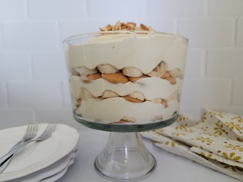 Trifle dish with layers of banana, nilla wafers, and pudding next to a stack of plates and forks