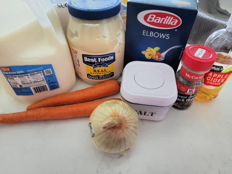 Milk, Best Foods Mayo, Elbow Macaroni, Carrots, Onion, Salt, Black Pepper, and Apple Cider Vinegar on a white counter. 