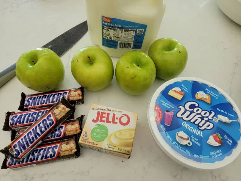 Snickers apple salad ingredients on a white counter, full size snickers bars, vanilla pudding, container of cool whip, 4 green apples, milk jug, and a sharp knife. 