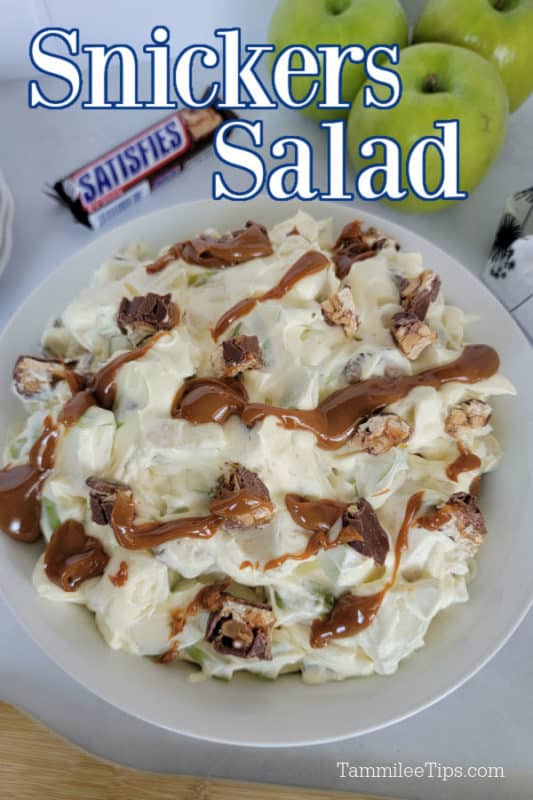 White bowl with snickers and apple salad covered in a bit of caramel, full size snickers bar and 3 green apples above the bowl. 