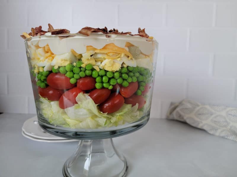 Traditional 7 layer salad in a trifle dish on a white counter with a cloth napkin behind. 