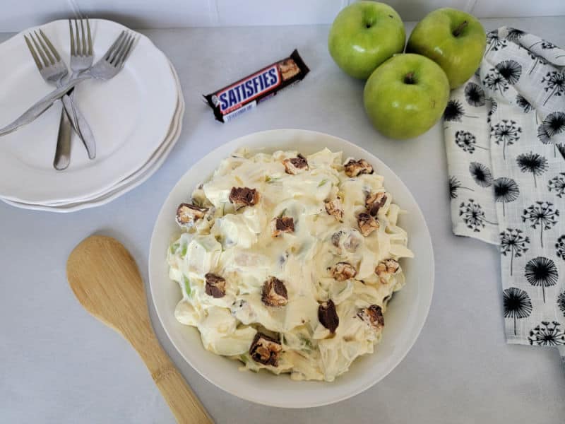 Snickers apple salad in a white bowl on a countertop with 3 apples, white plates with forks, and a Snickers Bar
