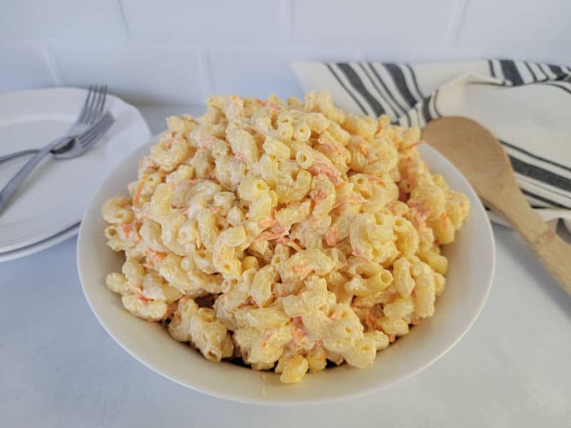 Macaroni salad with shredded carrots in a white bowl next to a wooden spoon, cloth napkin, and plates with forks. 