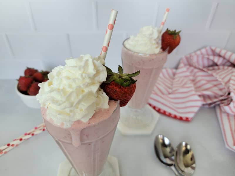 2 Strawberry milkshakes in glasses with a bowl of strawberries and 2 spoons. 