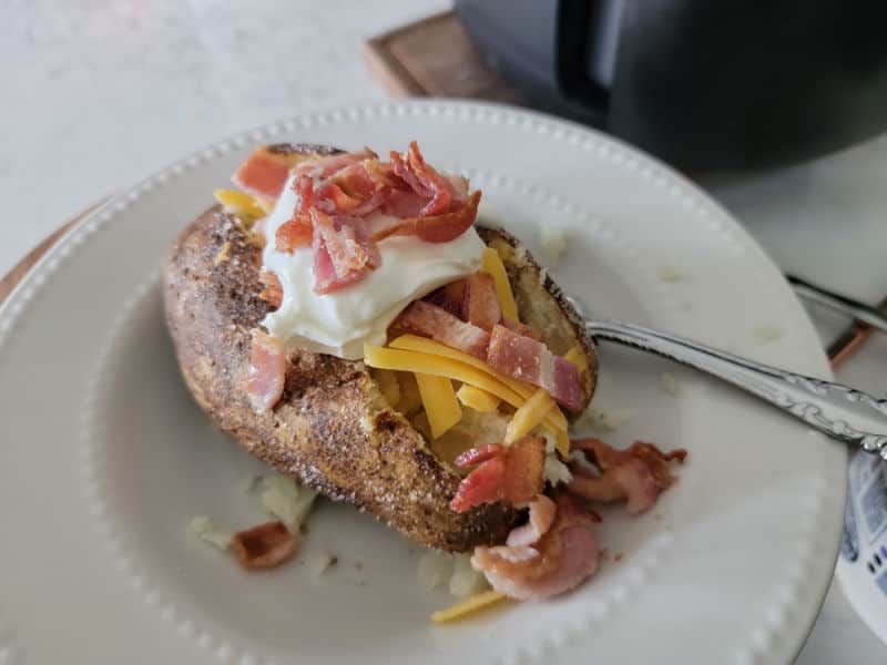 bacon, sour cream, cheese, on a baked potato on a white plate with a fork next to an air fryer 