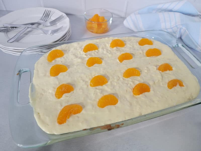 Pig pickin cake in a glass baking dish next to a bowl of mandarin oranges, a stack of plates and forks, and a blue cloth towel