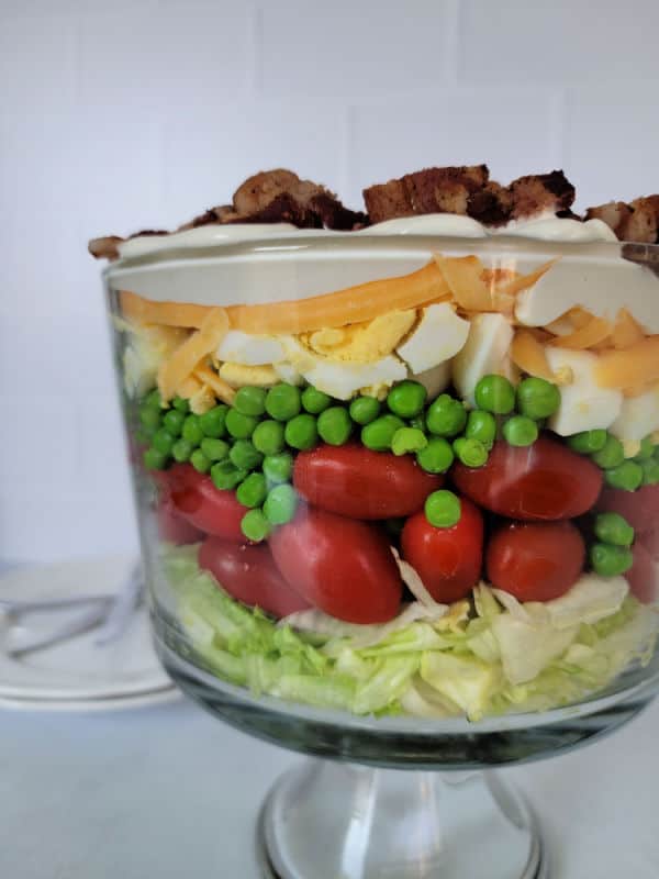 7 layer salad in a trifle dish with lettuce, tomato, peas, hard boiled eggs, dressing, cheese, and bacon. 