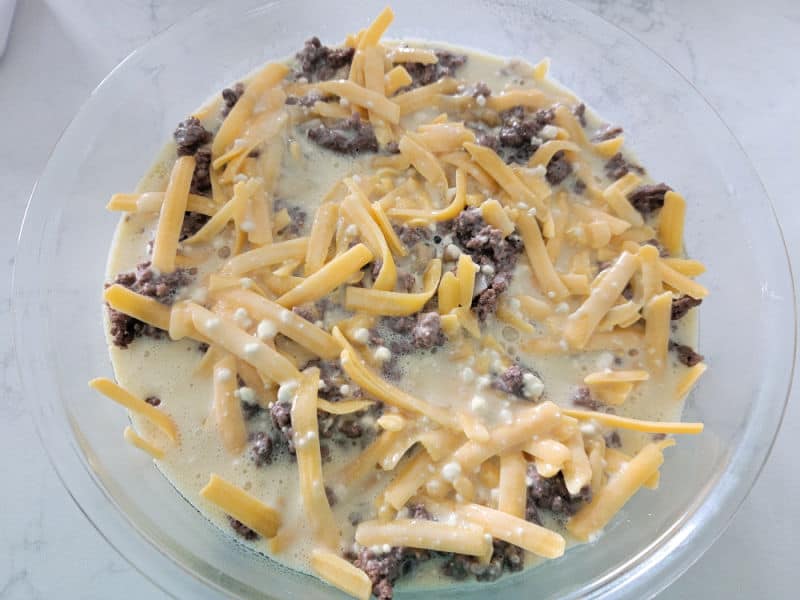 Bisquick Impossible Cheeseburger Pie with shredded cheddar cheese, ground beef, and bisquick mixture in a glass pie dish