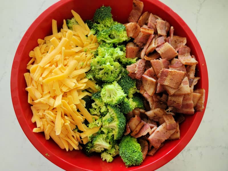Shredded cheddar cheese, broccoli florets, and bacon in a red bowl 