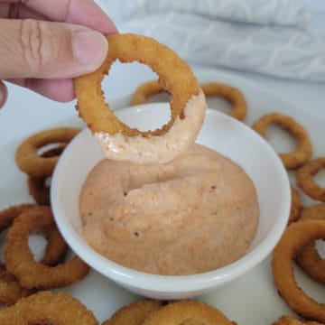 hand holding an onion ring dipping into a white bowl with bloomin onion sauce.