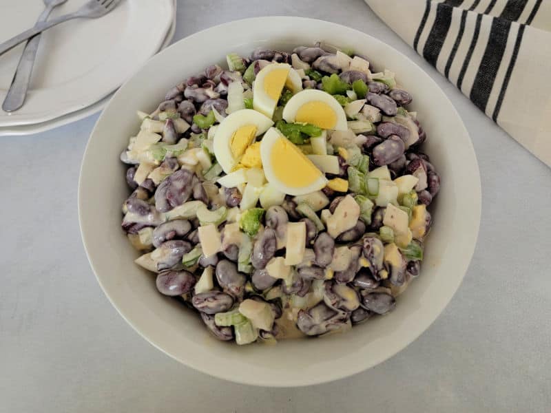 Kidney bean salad garnished with hard boiled egg slices in a white bowl next to plates with two forks and a cloth napkin