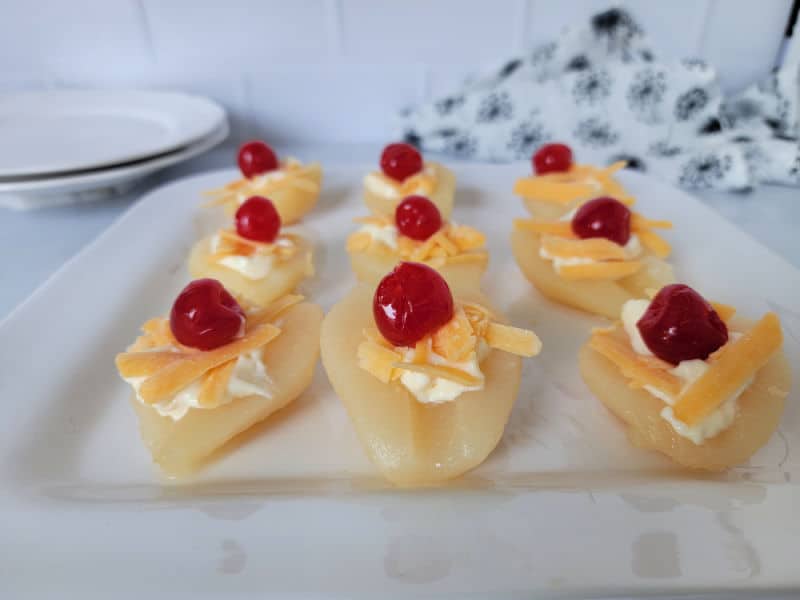 Pear halves with cheese and cherries lined on a white platter with plates in the background. 