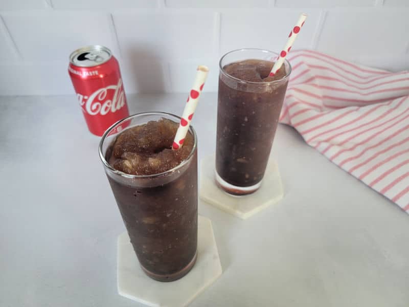 Two Coke slushies in tall glasses next to an open can of Coca Cola and a red striped cloth napkin
