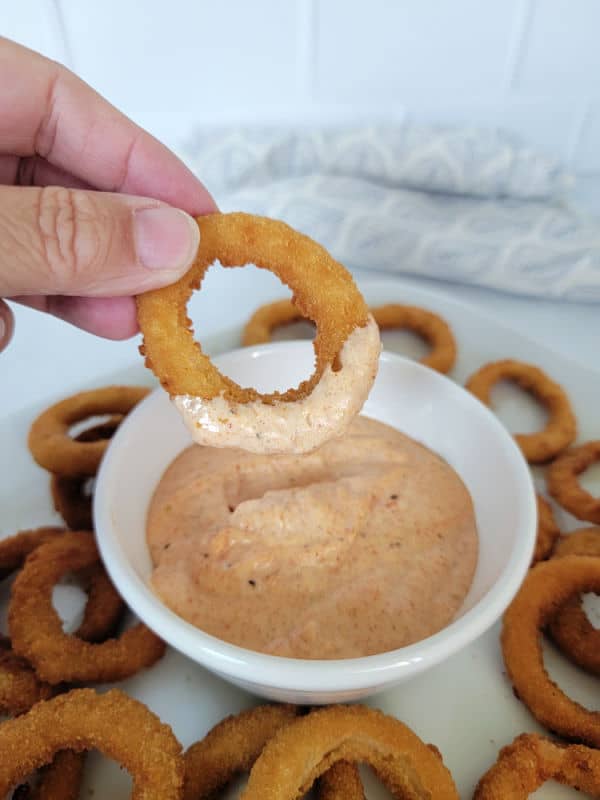 Hand holding an onion ring after it has been dipped in bloomin onion sauce in a white bowl