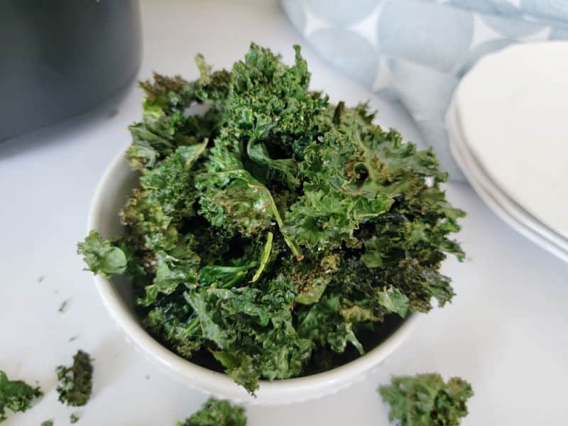 Kale chips in a white bowl next to plates and an air fryer