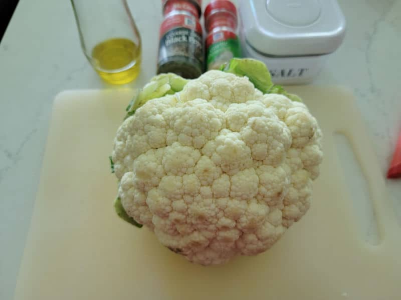 Cauliflower head on a white cutting board with salt, pepper, and garlic powder in the background. 