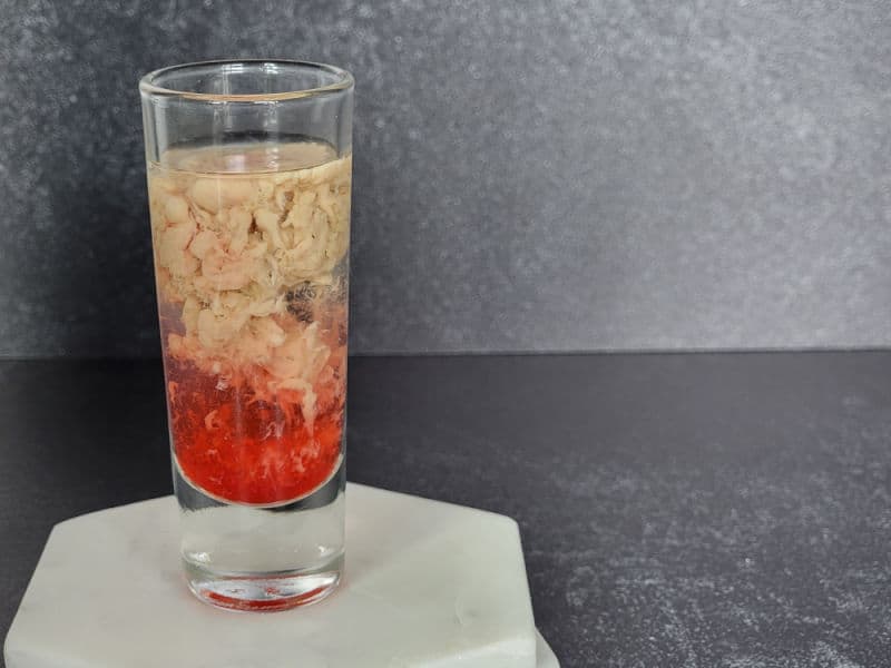 cream and red liquid swirling in clear liquid in a shot glass on a marble coaster