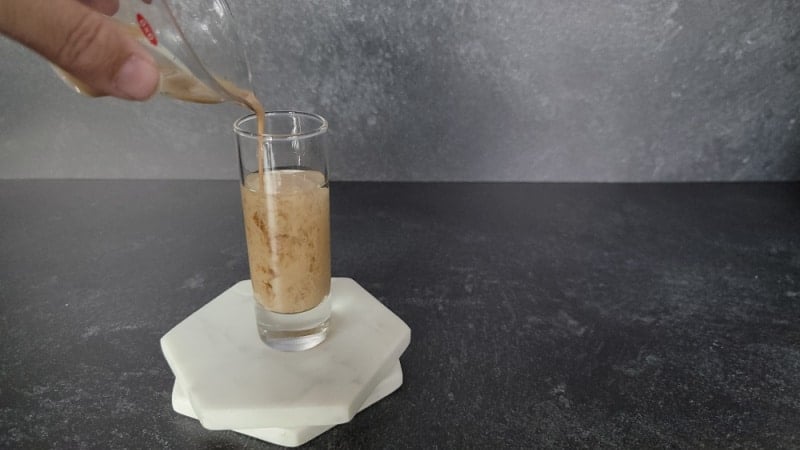cream liquid pouring into clear liquid in a shot glass on two marble coasters
