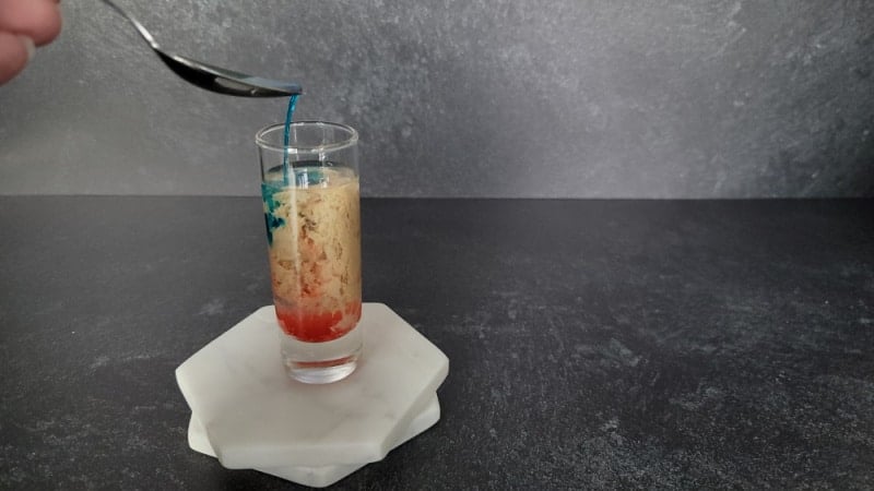 blue liquid pouring off a spoon into cream, red and clear liquid in a shot glass on two coasters. 