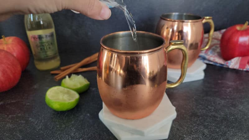 Clear liquid pouring into a copper mule mug on a coaster with apples, lime pieces, and ginger beer in the background. 