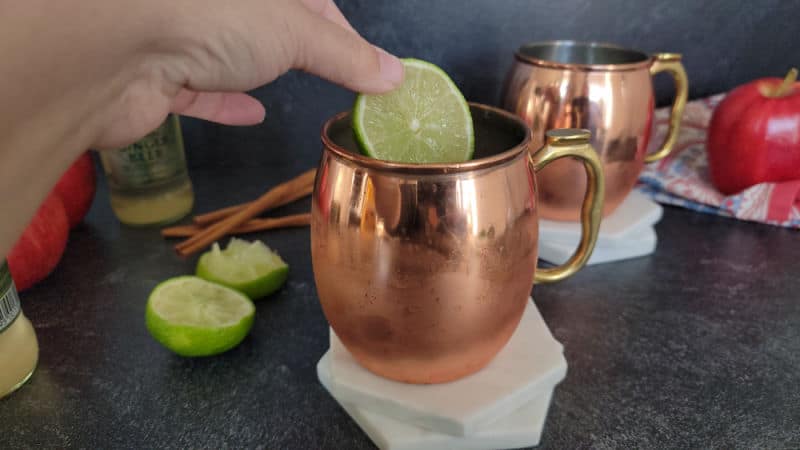 lime wheel being placed in a copper mule mug on two coasters, additional mug, apples and limes in the background