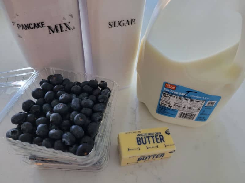 Pancake mix, sugar, fresh blueberries, butter, and milk on a white marble counter