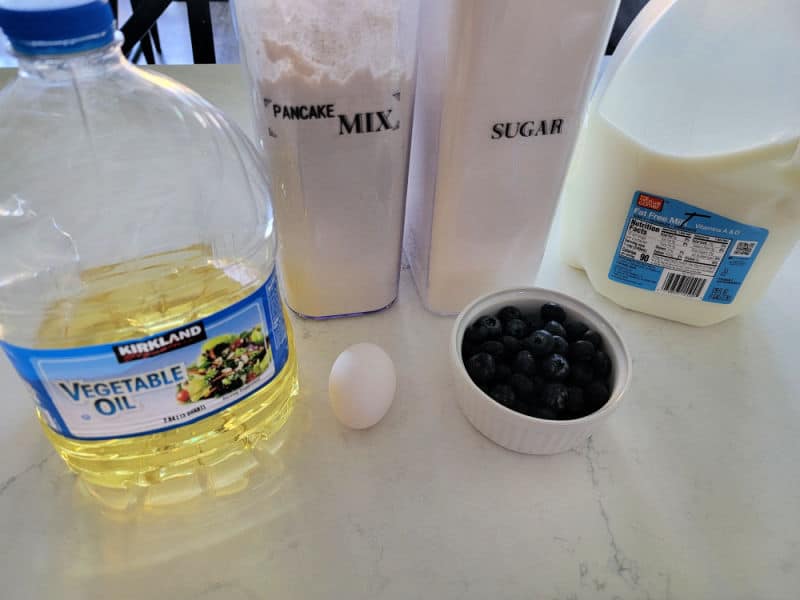 Vegetable oil, pancake mix, sugar, egg, blueberries, an milk on a white counter