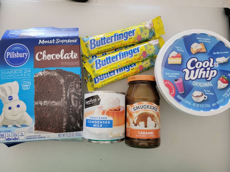 Butterfinger poke cake ingredients, chocolate cake mix, Butterfinger candy bars, cool whip, sweetened condensed milk, and caramel topping on a white counter