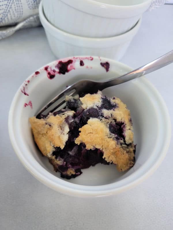 Small white bowl with blueberry cobbler and a fork next to a stack of bowls. 