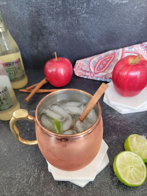 Copper mule mug with ice, lime, and cinnamon stick next to ginger beer, apples, cinnamon sticks, and lime wheels