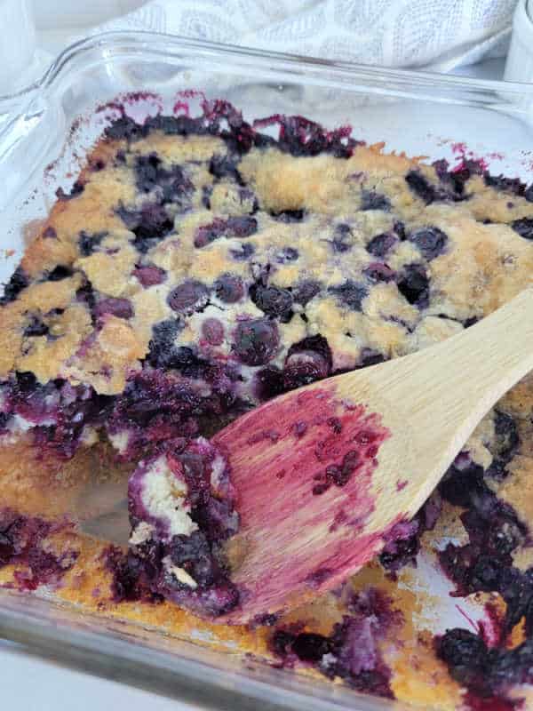 Wooden spoon covered in blueberry juice in a glass baking dish with blueberry cobbler. 