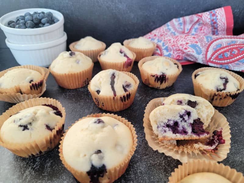 blueberry muffins on a dark counter next to a white bowl with fresh blueberries and a cloth napkin