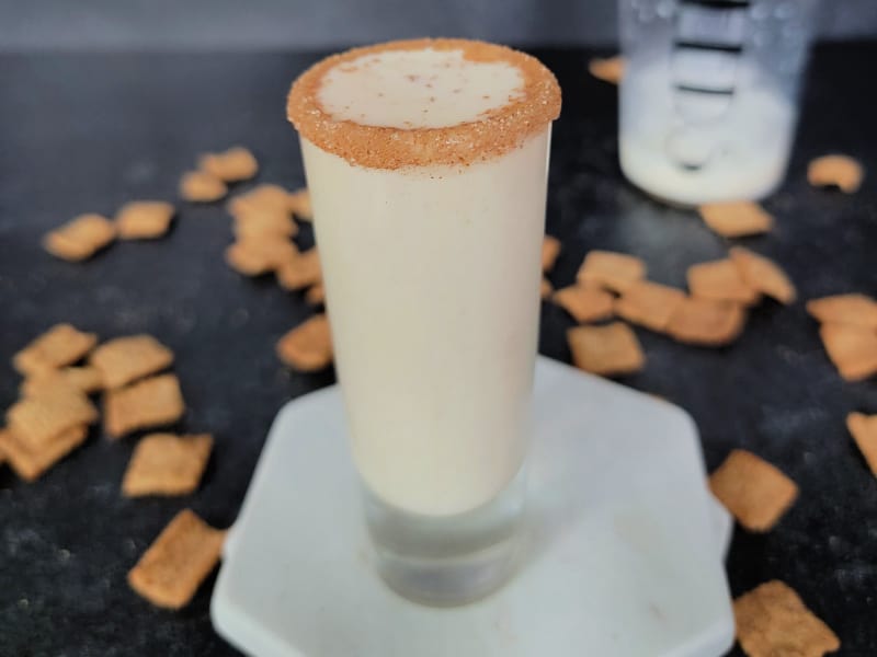 Cinnamon toast crunch shot on a white coaster surrounded by cereal and a cocktail shaker