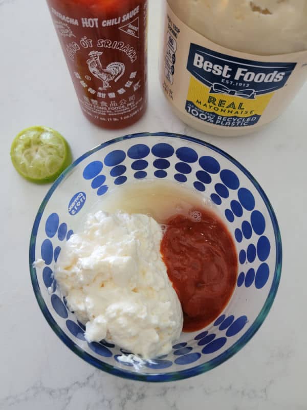 squeezed lime, bottle of sriracha, and bottle of best foods mayo behind a glass bowl with mayo, sriracha and lime juice in it. 