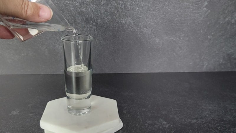 Clear liquid pouring into a glass shot glass sitting on two marble coasters. 
