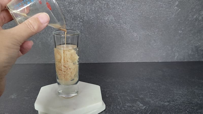 cream liquid pouring into the clear liquid in a glass shot glass sitting on two marble coasters 