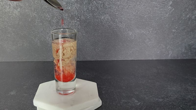 Red liquid dripping into a glass shot glass with cream and clear liquid in it