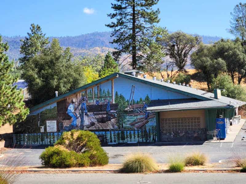 Logging mural on the side of a building with trees and mountains in the background