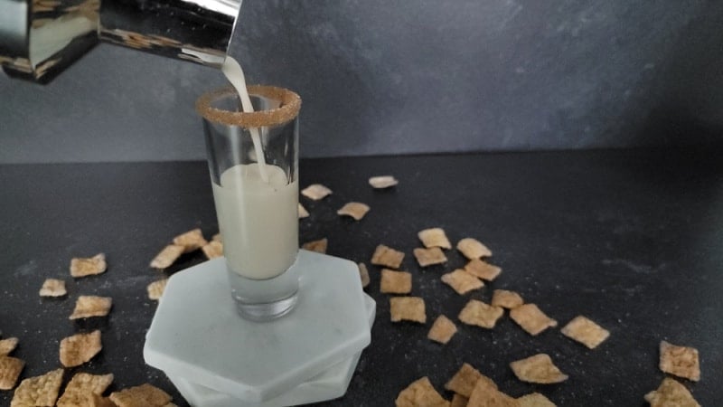 Cocktail shaker pouring into a shot glass on marble coasters with cinnamon toast crunch cereal spread around it