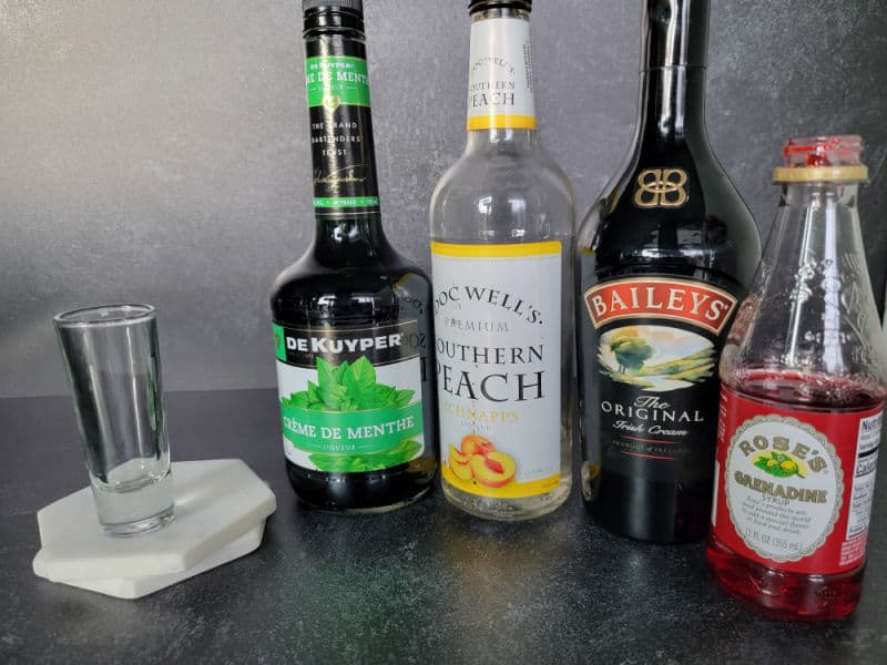 Shot glass next to bottles of creme de menthe, peach schnapps, baileys Irish cream, and grenadine