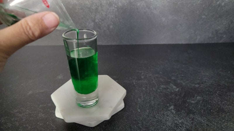 Green liquid pouring into shot glass on two marble coasters