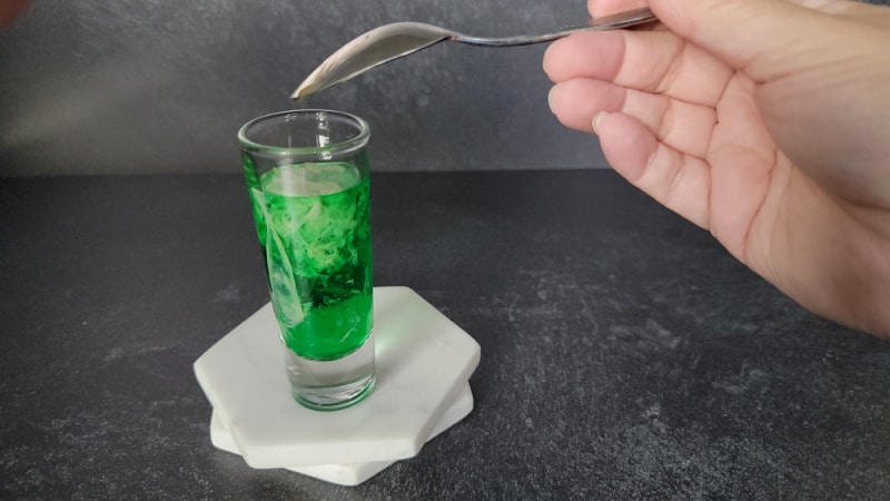 Spoon held upside down above a shot glass with green and cream liquid